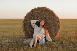 una joven a la hermosa luz de la puesta de sol de verano en un campo está sentada cerca de los fardos de paja. hermosa chica romántica con el pelo largo al aire libre en el campo foto