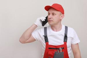 trabajador de la construcción en forma de trabajo roja y gorra con teléfono celular en la mano y se comunica con clientes aislados en fondo blanco. de la prestación de servicios es una carrera foto