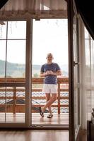 Young man standing on balcony of the hotel in the morning. holding a cup of coffee or tea in her hands. Looking outside nature forest and Mountain photo