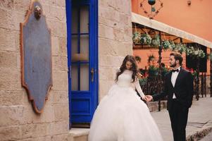 Wedding couple hugging in the old city. Blue vintage doors and cafe in ancient town on background. stylish bride in white long dress and groom in suit and bow tie. wedding day photo