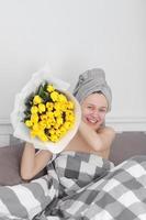 mujer feliz con flores de tulipán amarillo en la cama después de la ducha con una toalla en la cabeza disfrutando de un regalo romántico para el día de san valentín. le dio un ramo de flores. cumpleaños, día de la mujer, foto