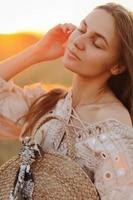 girl with long hair and straw bag in hand in the summer at sunset in the field for a walk. she is happy, her eyes are closed. background blurred art photography photo