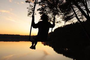 silhouette of a romantic young woman on a swing over lake at sunset. Young girl traveler sitting on the swing in beautiful nature, view on the lake photo