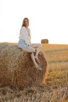 Young woman with long hair, wearing jeans skirt, light shirt and straw bag in hand, sitting on bale on field in summer. Female portrait in natural rural scene. Environmental eco tourism concept. photo