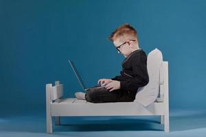 schoolboy in glasses using laptop sitting on bed. Child doing school homework. Online learning, distance lesson, education with online tutor at home isolation photo