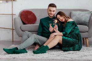 happy couple in stylish green clothes with sparklers. Man and pregnant woman sitting near sofa. Valentine's Day photo