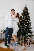 Christmas family photo with christmas tree and ornaments, wooden floor and fireplace. Man and woman are kissing near christmas tree.