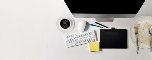 escritorio de oficina blanco moderno con computadora de pantalla negra, una taza de café, un cuaderno, un bolígrafo, un plato de comida que se ha comido. vista superior con espacio de copia, tumbado foto