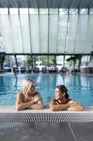 Shot of two women cheering with drinks near swim pool. Red, green and yellow different cold cocktails with ice cubes, straws, transparent clean blue aqua in pool photo