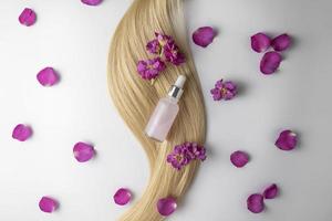 A hair care oil or serum in a dropper bottle lying on a blond hair strand with flowers and rose petals around it, product marketing mockup photo