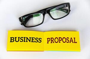 Top view of business proposal text on yellow notepad with glasses on white cover background. Business concept photo