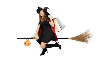 Beautiful girl with black and orange hair  in black dress and witch hat is riding staff with shopping bags on white background for Halloween day.Emotion smile photo
