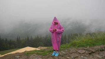 mujer joven en poncho de lluvia rosa se encuentra en la cornisa con vistas a un bosque de niebla video