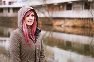 mujer joven perforada con cabello rosado y parka encapuchada de pie junto al río foto