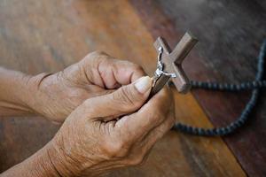 Closeup wooden cross which has a metal statue of crucified Jesus is in the hands of an asian eldery Catholic while praying in a local church, soft and selective focus. photo