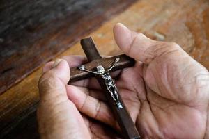 Closeup wooden cross which has a metal statue of crucified Jesus is in the hands of an asian eldery Catholic while praying in a local church, soft and selective focus. photo
