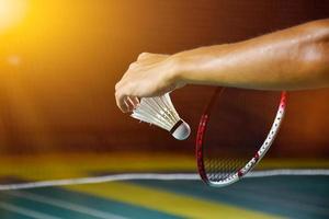 la raqueta de bádminton y el viejo volante blanco sosteniéndose en las manos del jugador mientras lo sirven sobre la red por delante, desdibujan el fondo de la cancha de bádminton y el enfoque selectivo. foto