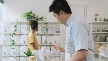Asiatischer männlicher Ladenbesitzer arbeitet mit Laptop, prüft Bestellung, ein Kunde wählt natürliches Bio-Produkt in den Ausstellungsregalen im Nachfüllladen, umweltfreundlicher und plastikfreier Laden, wiederverwendbarer Behälter. video
