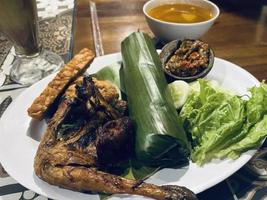 Indonesian cuisine or sundanese food called Nasi Timbel, Rice wrapped in banana leaves, served with fried chicken, tempeh, tofu, salted fish, chili sauce, lalap and sayur asem. photo