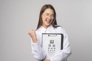 Young female ophthalmologist with glasses holding eye chart over blue background studio, healthcare concept photo