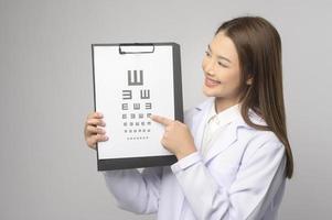 Young female ophthalmologist with glasses holding eye chart over blue background studio, healthcare concept photo