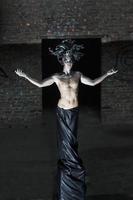 man on a pedestal with a naked torso and a headdress in the form of a bunch of snakes among the ruins and a window in the ceiling photo