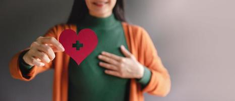 Love, Health Care, Donation and Charity Concept. Close up of Smiling Volunteer Woman Holding a Heart Shape with Cross Sign Paper. presenting to Camera photo