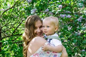 Candid portrait of young smiling mother with her little son in garden with blooming lilac flowers. photo