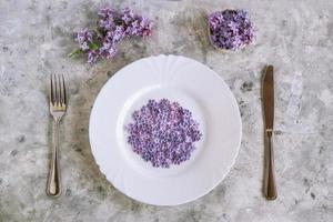 Served plate with purple lilac flowers with five petals, fork and knife, branch and bouquet of lilac flowers on gray concrete background, top view. photo