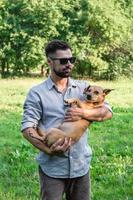 Handsome stylish European man is holding his dog in hands in park on a walk. Friendship between human and pets. photo