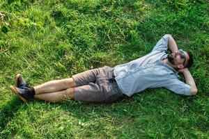 joven y guapo deportista europeo con gafas de sol descansa sobre un césped en el parque de verano, vista superior. foto