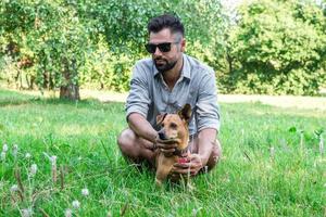 Handsome stylish European man sitting on grass with his dog in park on a walk. Friendship between human and pets. photo