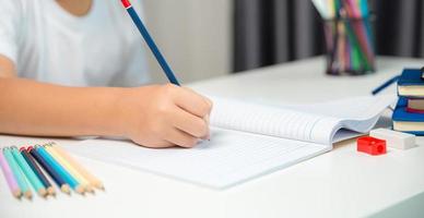 niño sentado estudiando en casa, niño estudiante en línea aprendiendo y haciendo deberes en el escritorio, niño pequeño leyendo y escribiendo un libro en la mesa. concepto de educación, ciberespacio tecnológico. foto