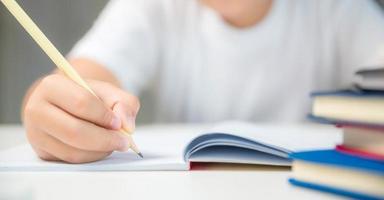 niño sentado estudiando en casa, niño estudiante en línea aprendiendo y haciendo deberes en el escritorio, niño pequeño leyendo y escribiendo un libro en la mesa. concepto de educación, ciberespacio tecnológico. foto