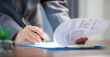 Workplace closeup person professional businesswoman sitting at desk hold pen signing or signature contract paper. Employee woman writing agreement document on paperwork form corporate at work office photo