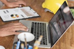 un ingeniero civil trabaja en su computadora portátil en su escritorio con un casco amarillo en su oficina en un sitio de construcción. foto