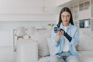 Satisfied young disabled girl install her modern bionic prosthetic arm, sitting on sofa at home photo