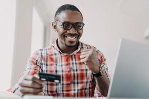hombre afroamericano que usa el servicio de banca en línea en una computadora portátil, con tarjeta de crédito hace un gesto de sí foto