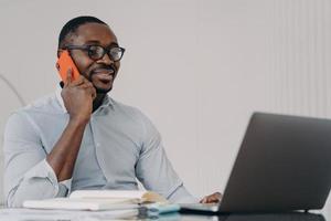 African american businessman in glasses makes business call, talking on phone, working at laptop photo