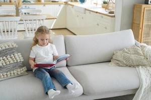 Small girl preschool child reads fairy tale stories holding paper book sitting on sofa alone at home photo