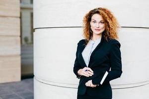 Horizontal portrait of serious pretty businesswoman with curly hair, thin eyebrows and curly hair, wearing black suit and white blouse, holding notebook with pen in hands, looking directly into camera photo