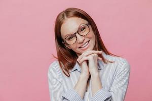 Elegant pretty woman tilts head, keeps hands under chin, wears transparent glasses and shirt, smiles gently, models over pink background. People and positive emotions concept. Charming female photo