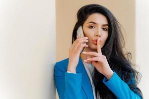 Mysterious brunette woman with makeup, talks with business party, tells secret, makes silent gesture, looks straightly at camera, wears formal apparel. People, communication and conspiracy concept photo