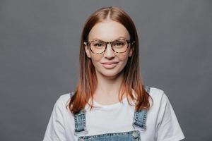 Happy young woman with glad facial expression, has healthy skin, wears optical glasses, white t shirt and denim sarafan, models over grey background, going to meet with friends, poses indoor photo