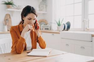 Female freelance worker making phone call, discussing work project in kitchen at home. Remote job