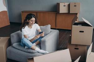 Smiling girl holding smartphone, selects moving company online for relocation, sitting with boxes photo