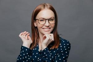 Positive attractive Caucasian woman keeps hand near chin, looks positively at camera, holds hand under chin, smiles happily, wears black shirt with white polks dot, poses over grey background. photo