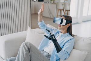 Modern disabled girl in virtual reality glasses playing video game, learning to use prosthetic arm photo