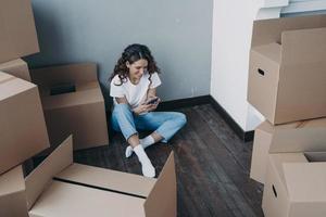 Modern girl sits with boxes uses smartphone app, choosing moving company and mover for relocation photo