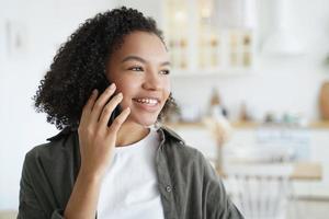 feliz niña de raza mixta responde llamadas, habla por teléfono, disfruta de una agradable conversación móvil en casa